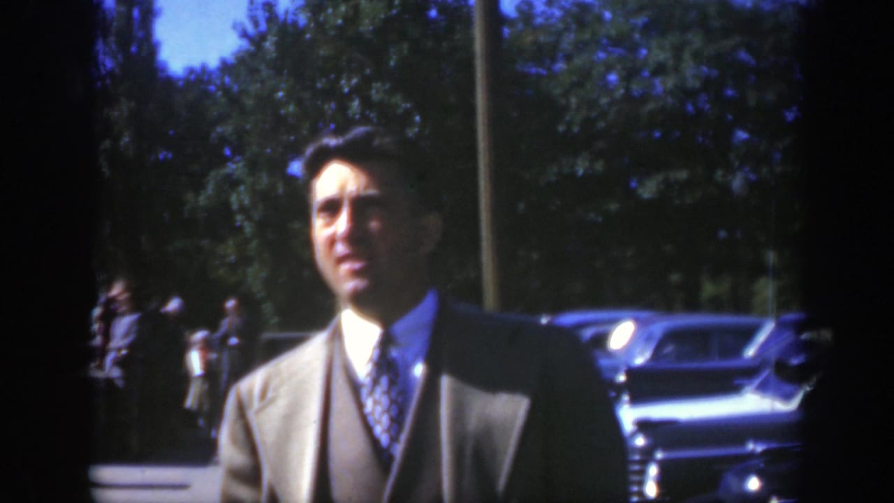 In Montreal circa 1950, the suited man looks toward the camera while vintage automobiles and pedestrians appear behind him in bright daylight.