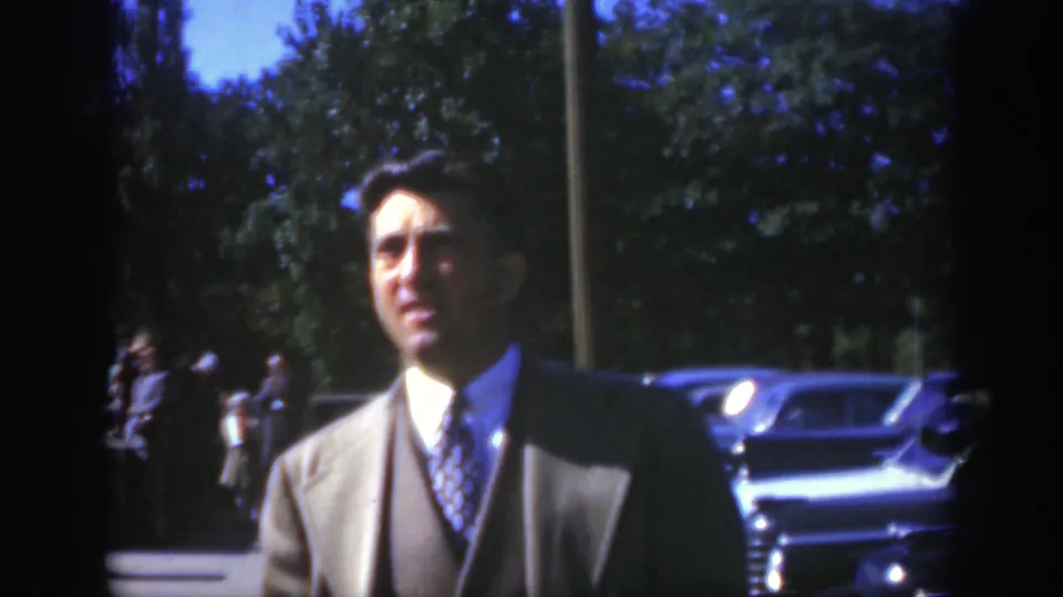 In Montreal circa 1950, the suited man looks toward the camera while vintage automobiles and pedestrians appear behind him in bright daylight.