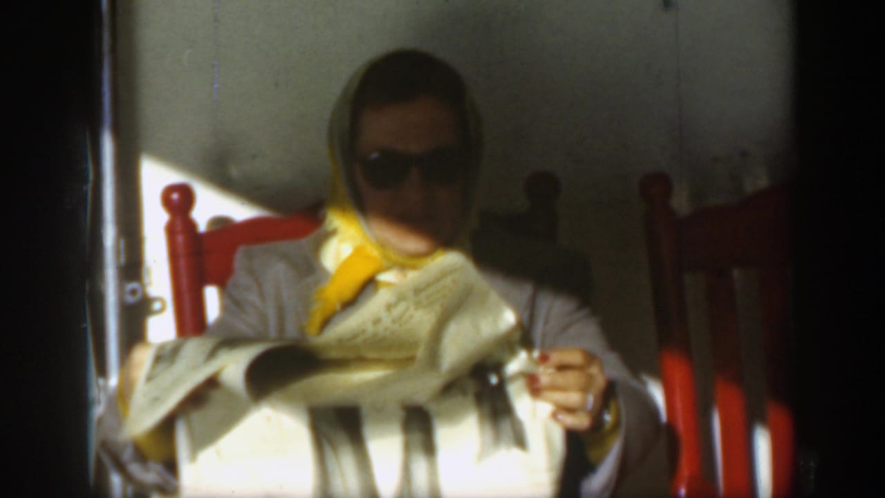 A woman in sunglasses and a yellow headscarf sits in a red chair reading or holding a newspaper, consistent with the circa-1950 home-movie scene.