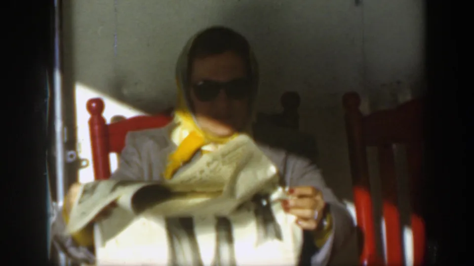 A woman in sunglasses and a yellow headscarf sits in a red chair reading or holding a newspaper, consistent with the circa-1950 home-movie scene.