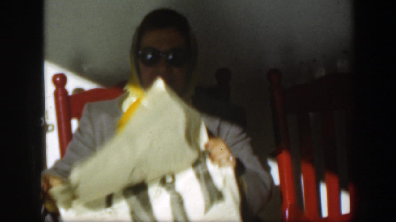 The seated woman in sunglasses and a yellow headscarf lifts or turns a newspaper while red chairs and a shaded wall remain visible behind her.