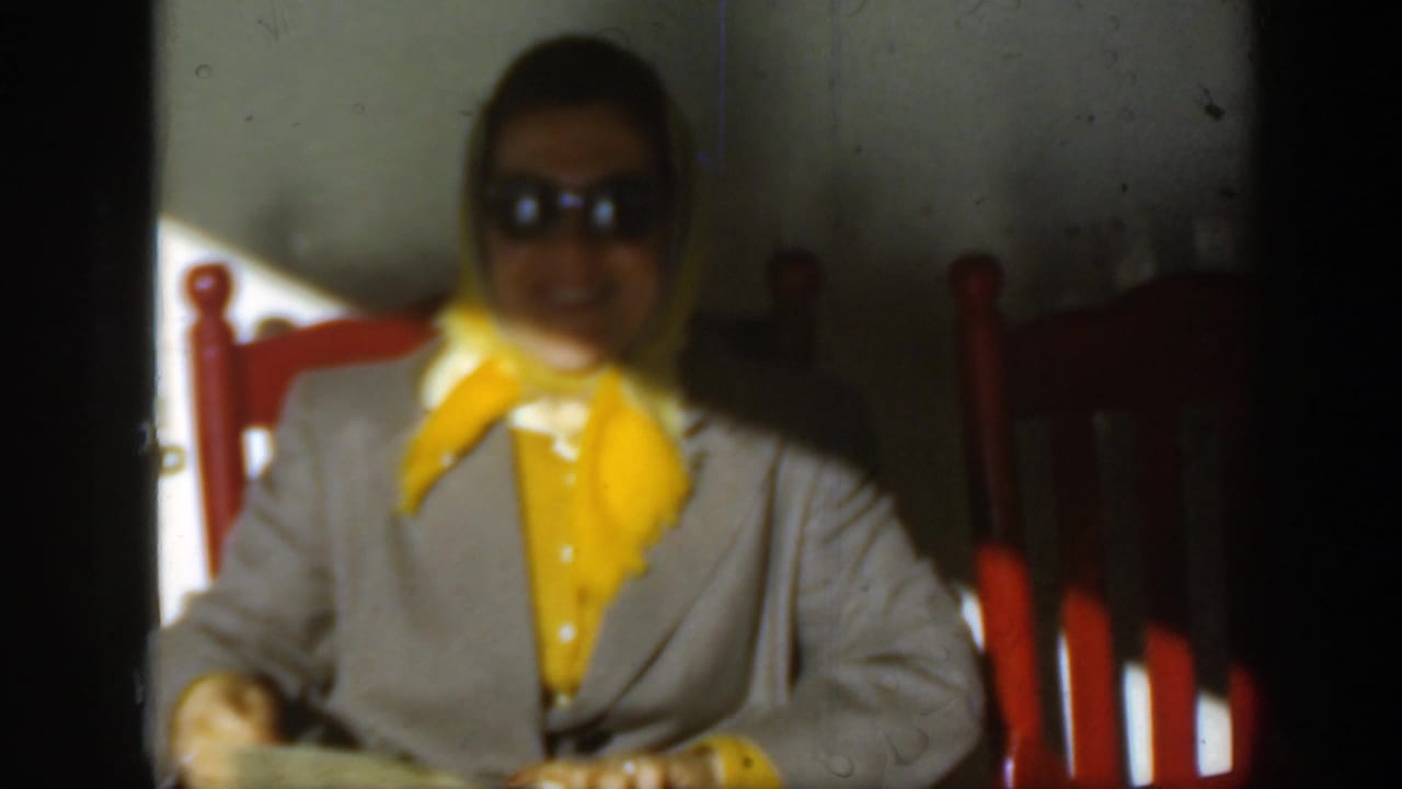 Woman in sunglasses and yellow scarf seated in a red wooden chair.