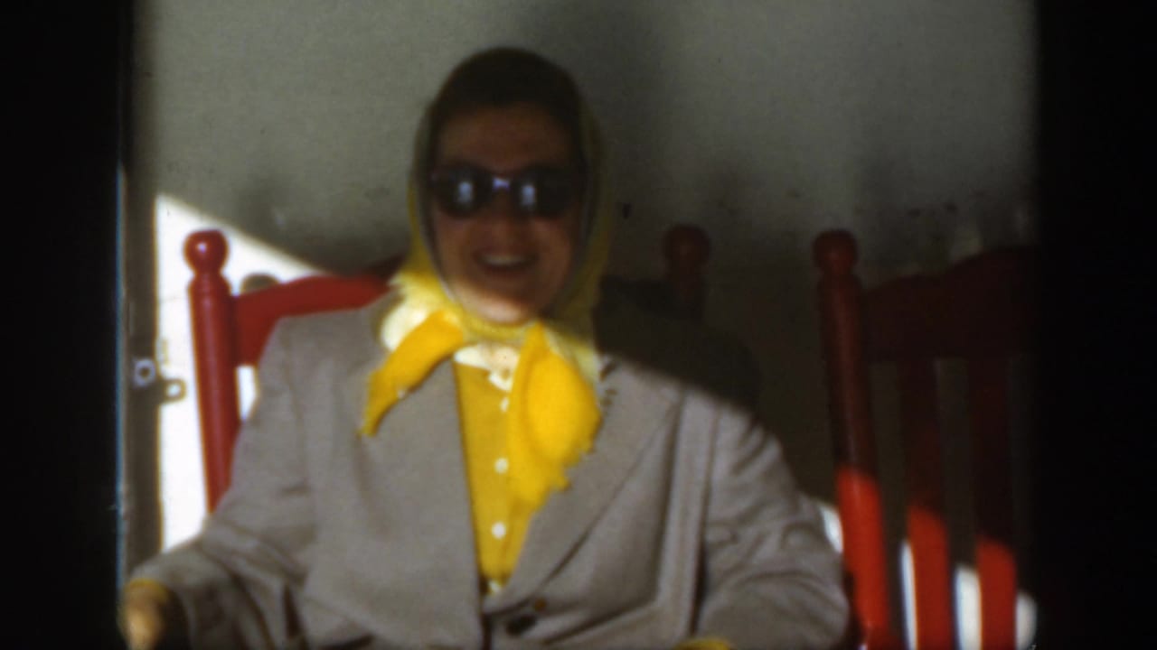 Smiling woman in sunglasses sits against a wall beside red chair backs.