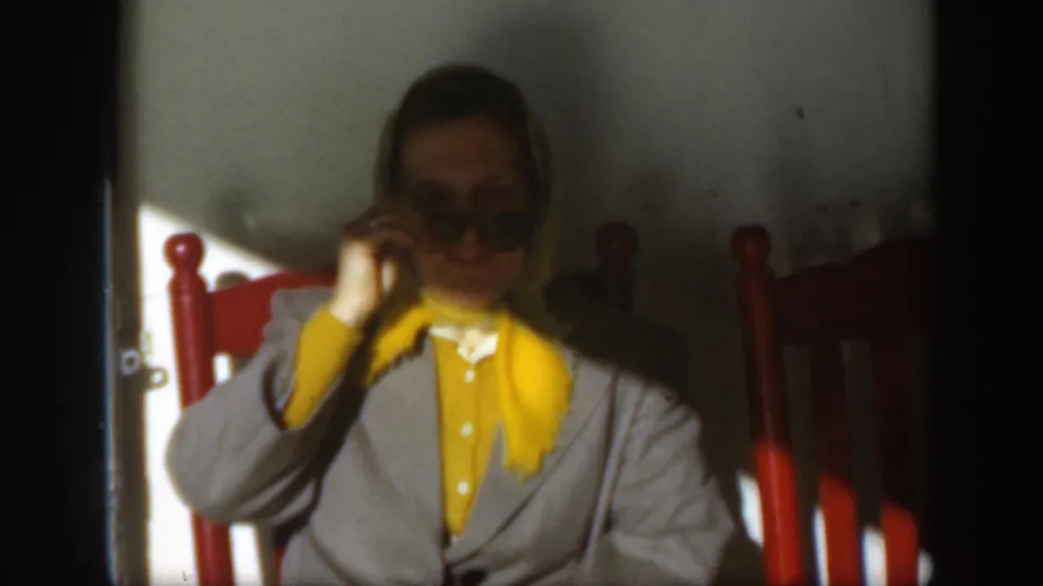 The woman raises one hand to adjust or remove her dark sunglasses while seated in the red chair.
