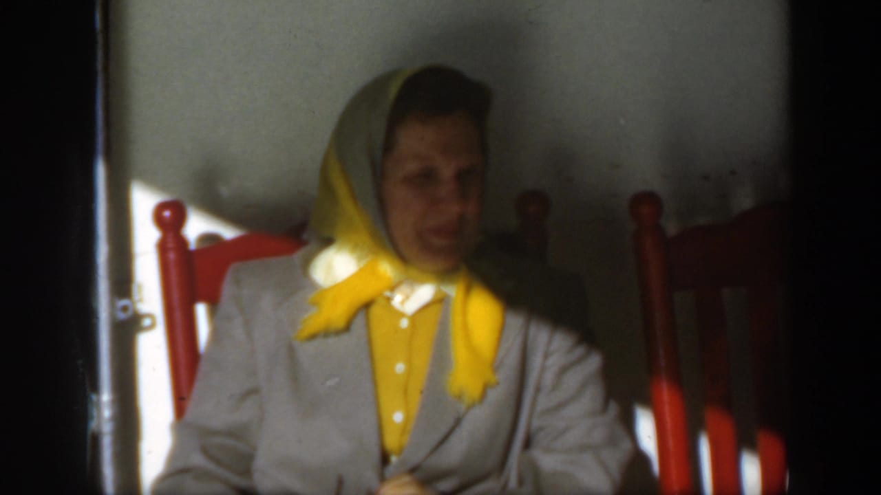 Woman in a gray suit and yellow headscarf seated on a red porch chair.