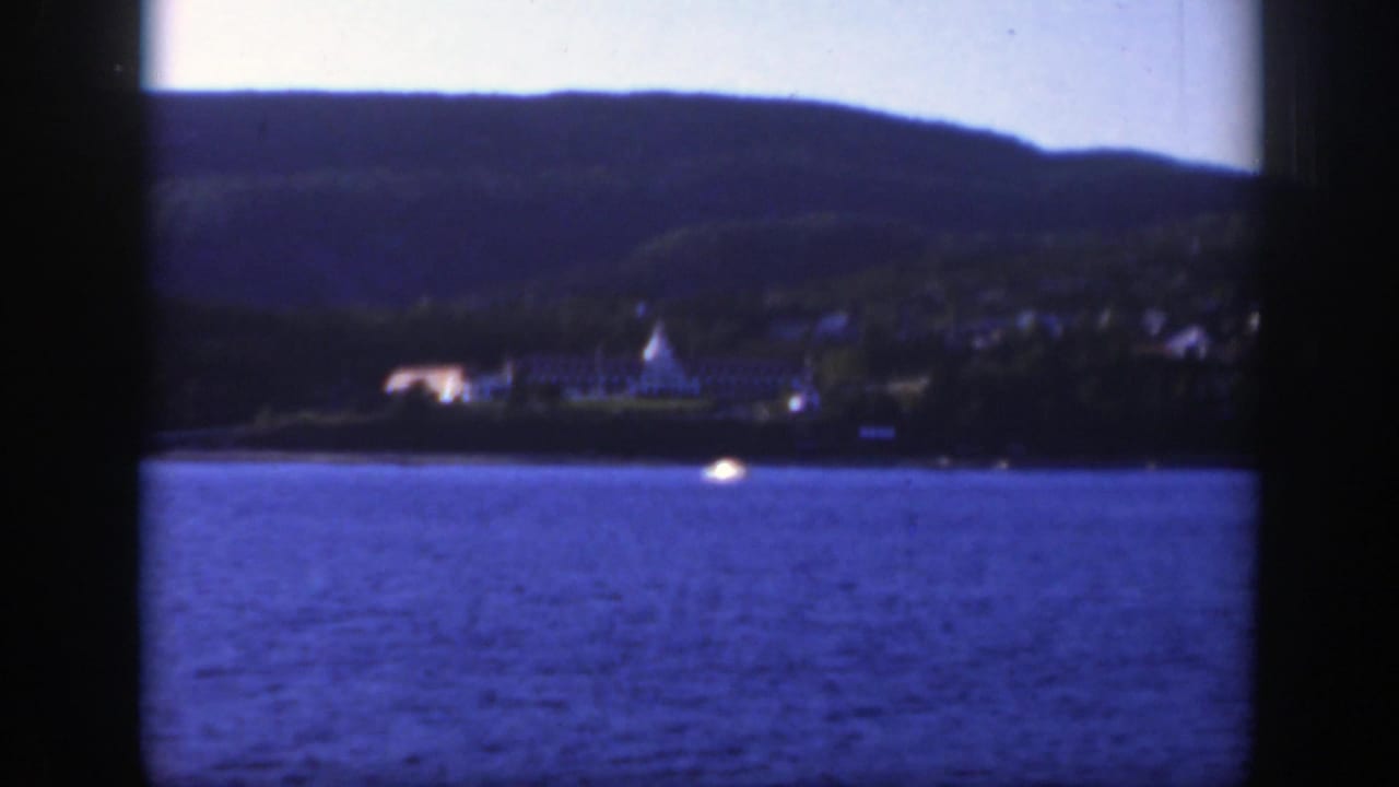 1950 Montreal-area 8mm frame looking across blue water toward distant lakeside buildings and tree-covered hills.