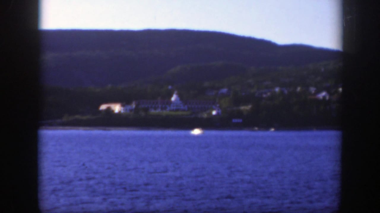 1950 8mm view toward a Montreal shoreline across a wide river, with a pale building and dark wooded hills in the distance.