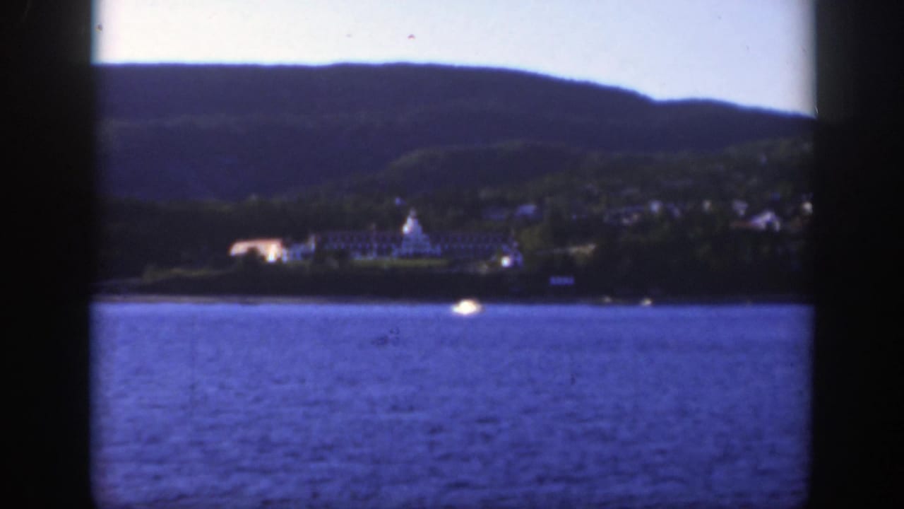 Soft 8mm frame of Montreal's distant riverfront, with a large pale structure, hillside trees, and a small boat on the water.