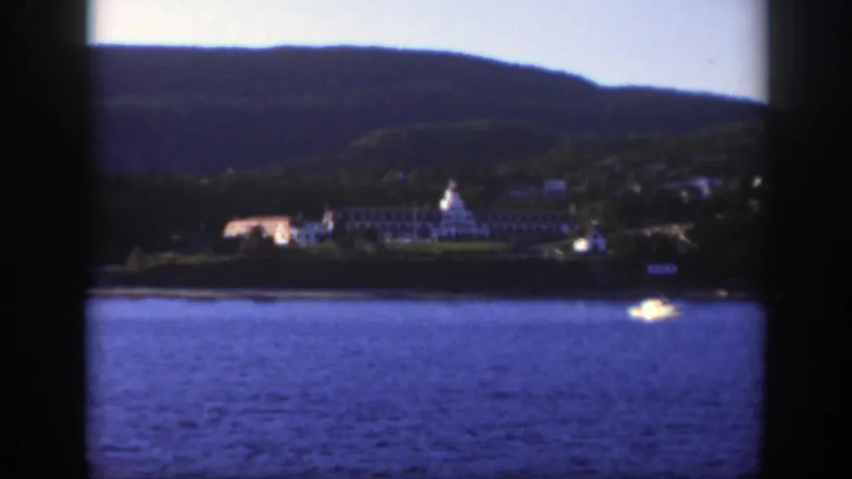 1950 home-movie frame showing a broad river, a distant pale shoreline building, small boats, and forested hills near Montreal.