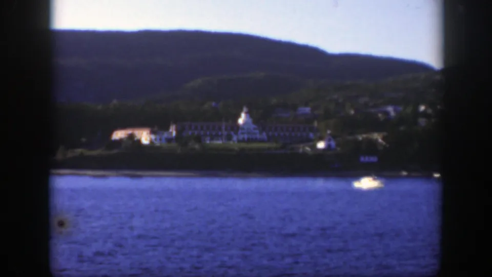 Distant Montreal river scene on 8mm film, with a bright boat crossing the water and shoreline buildings under wooded hills.