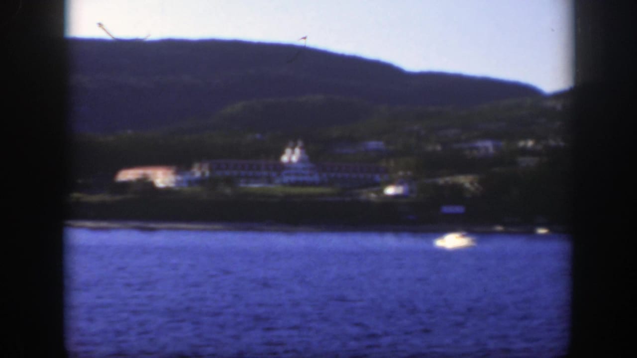 1950 8mm view near Montreal showing a white lakeside resort across blue water with wooded hills and a small boat in the foreground.