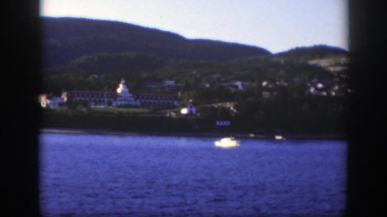 1950 8mm frame near Montreal with a white lakeside hotel, green hillside, blue water, and a small bright boat near the shore.