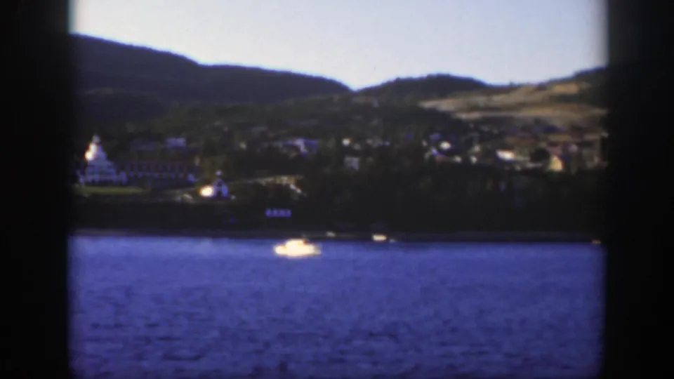 1950 8mm frame near Montreal showing a white resort on the shore, wooded hills, open blue water, and a small light-colored boat.