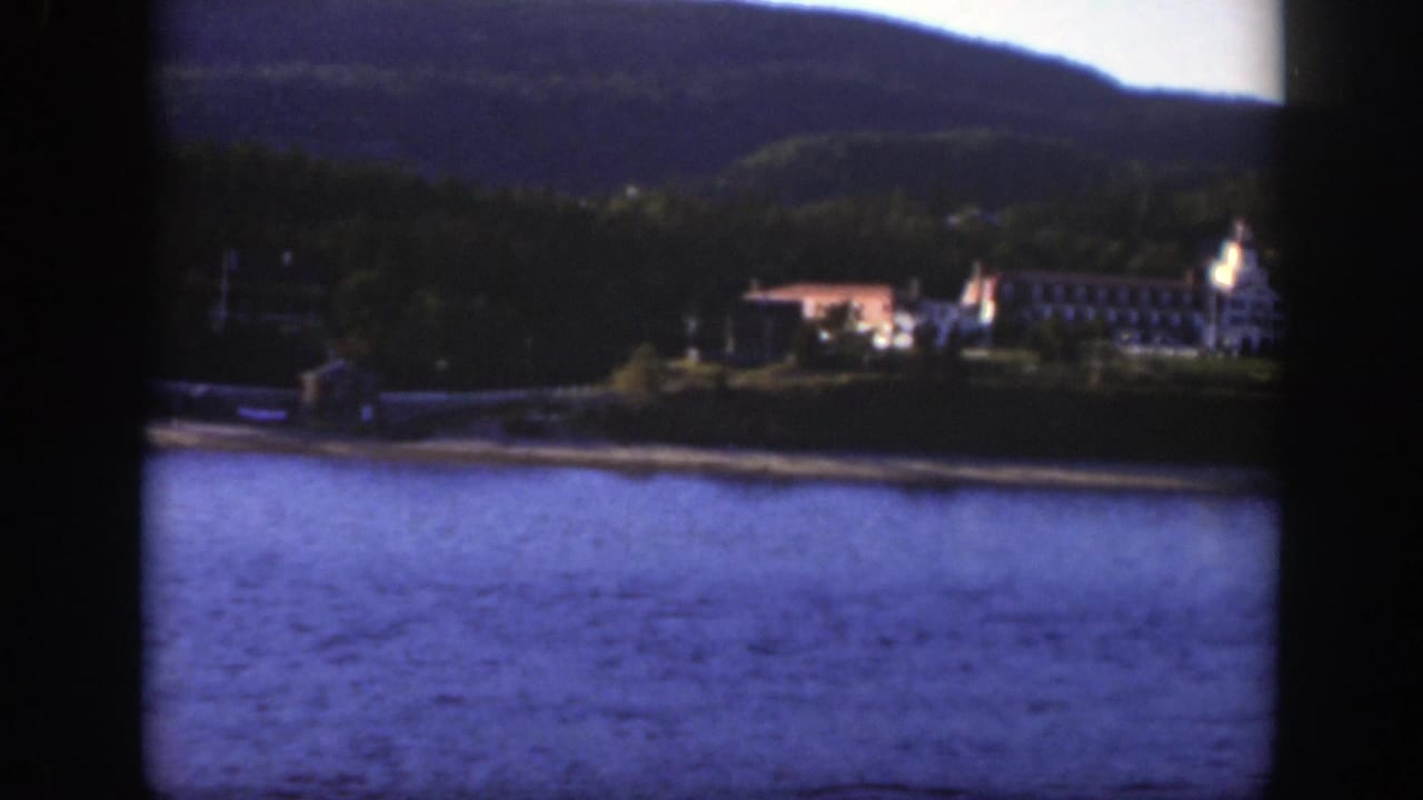 1950 Montreal-area home-movie view of a large shoreline building complex across blue water, backed by trees and hills.