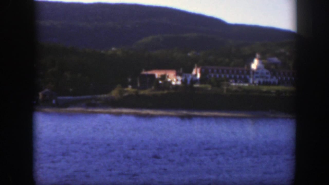 A steady 1950 home-movie frame shows white buildings along a Canadian lakeshore with forested hills in the background.