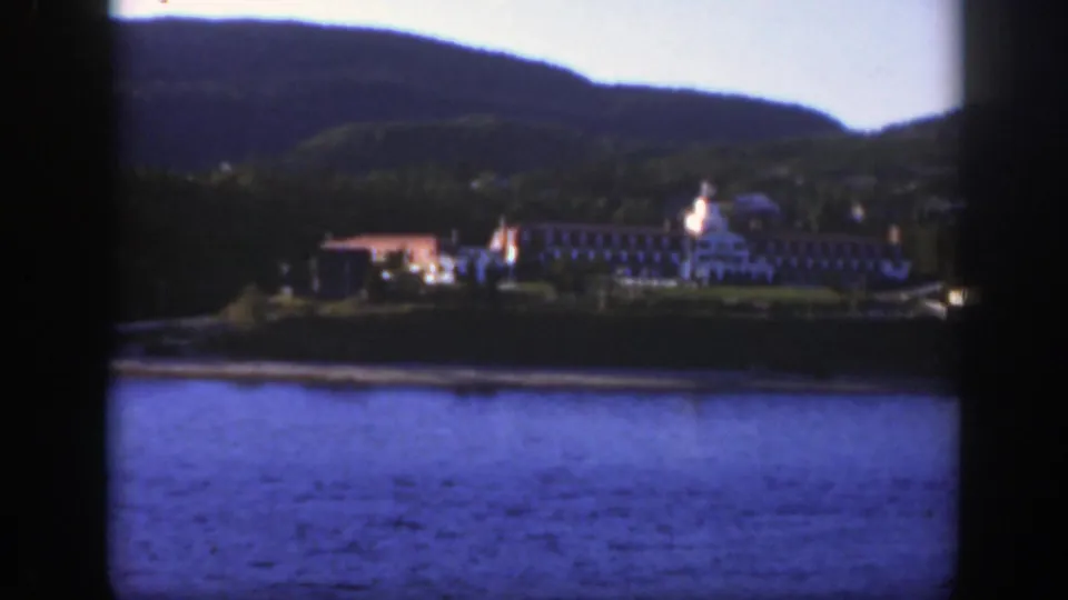 The camera holds on a broad lakeside complex near Montreal, with red-roofed structures, trees, water, and hills visible.