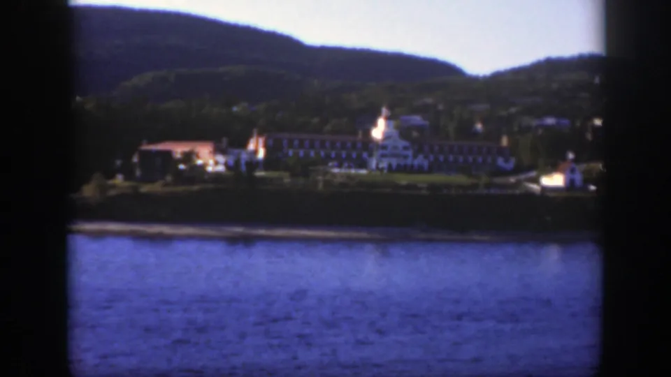 A 1950 home-movie view shows the lakeside complex, wooded hills, blue water, and a small white vessel near the right shoreline.