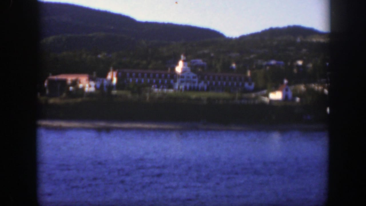 1950 8mm view near Montreal of a large lakeside resort seen across water, with wooded hills behind and dark framing in the foreground.