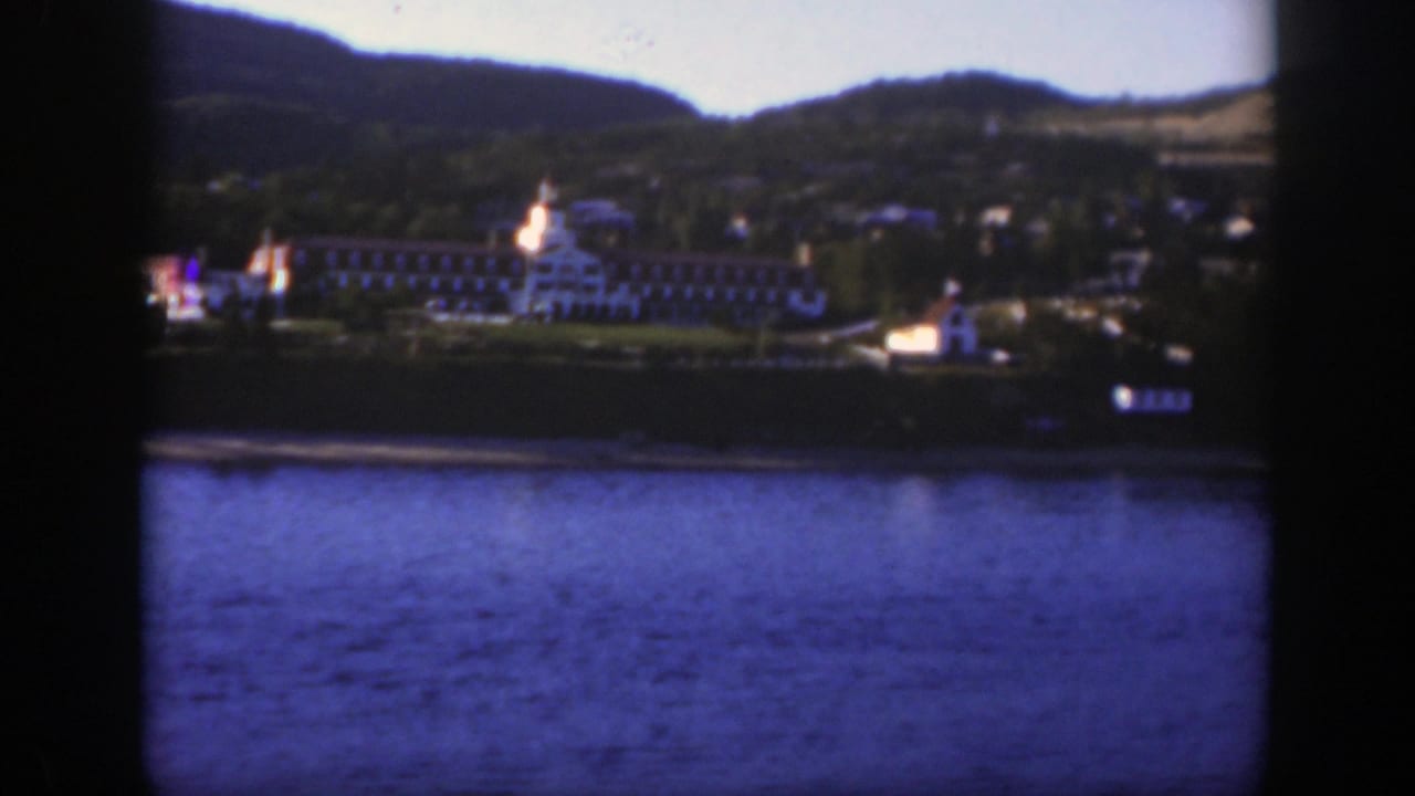 A distant resort complex along a calm lake near Montreal, with a prominent white building, shoreline structures, and hills in the background.