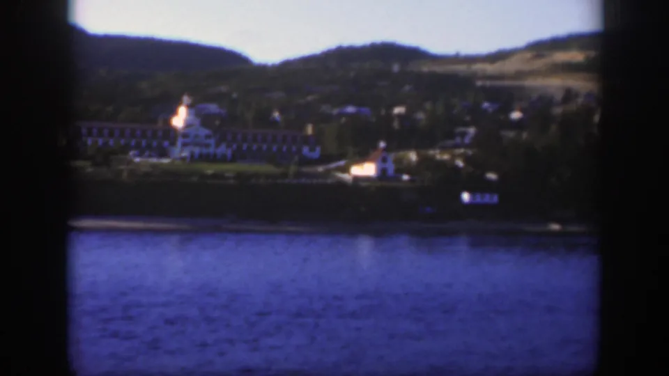The 1950 home-movie frame shows the resort shifted left across the lake, with a bright shoreline structure and rolling hills behind.