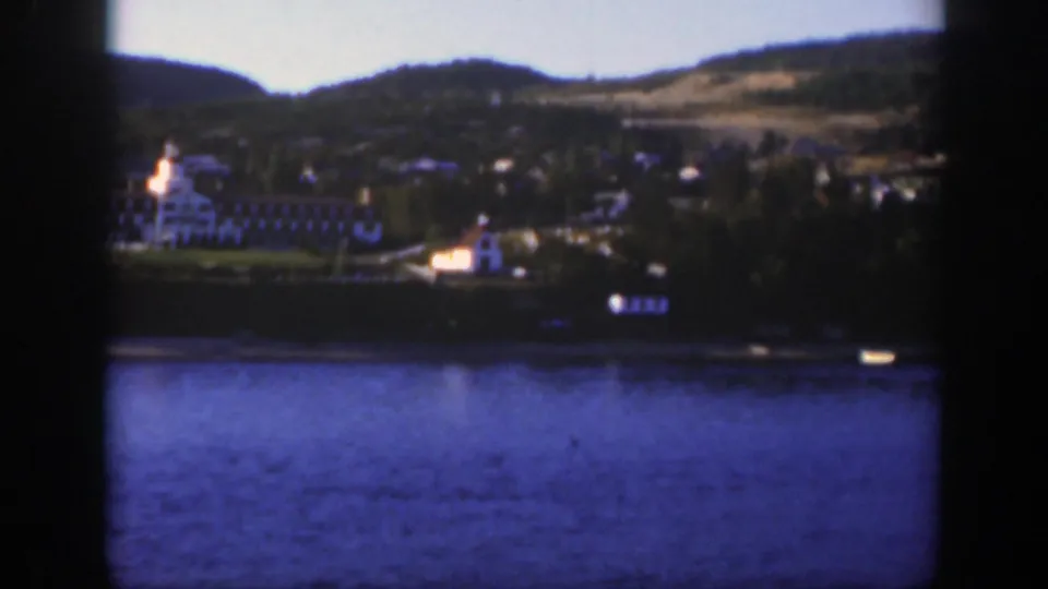 A distant Montreal-area resort across blue lake water, framed by dark vertical bars with hills and scattered buildings behind.