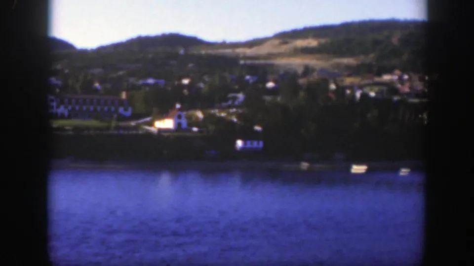 1950 Montreal-area 8mm frame showing blue lake water, a distant white shoreline building and wooded hills.