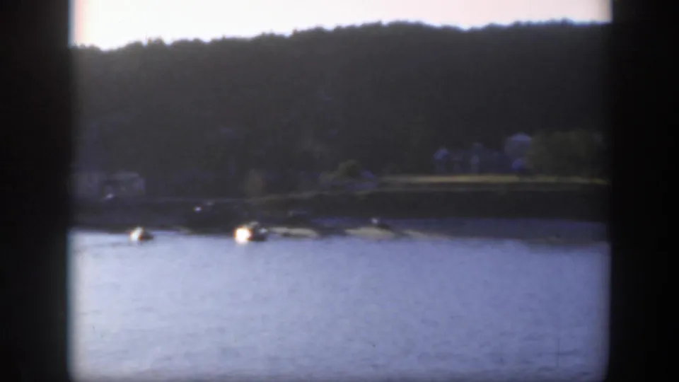 1950 Montreal-area lakeside frame showing bright water, small boats near shore and a dark wooded hill in the background.