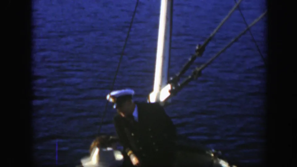 A man in a dark uniform and white cap stands on a boat deck with rigging and blue water behind him.