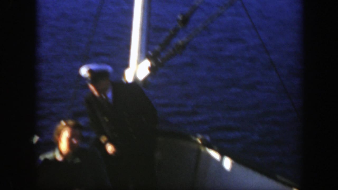 1950 Montreal home-movie frame showing a white-capped captain on a boat deck in low light beside dark blue water.