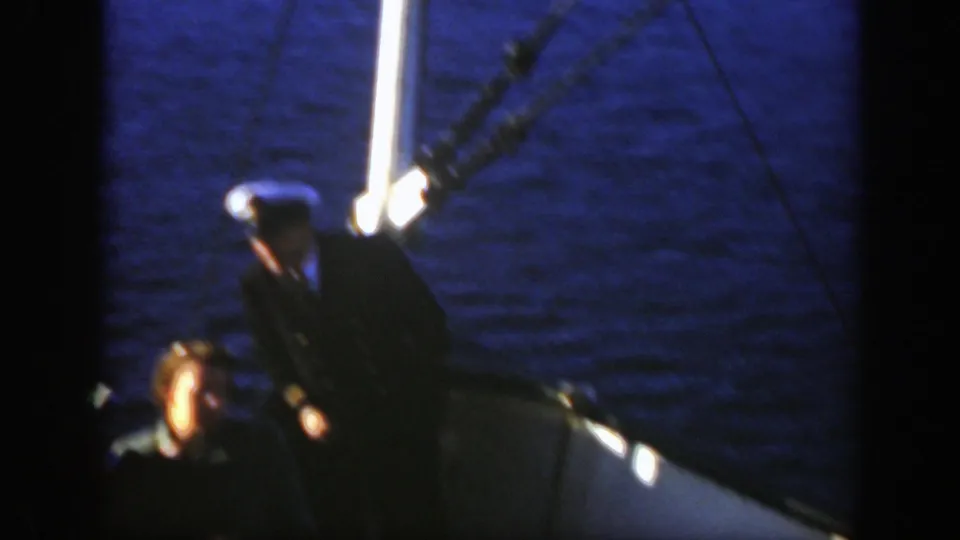 1950 Montreal home-movie frame showing a white-capped captain on a boat deck in low light beside dark blue water.