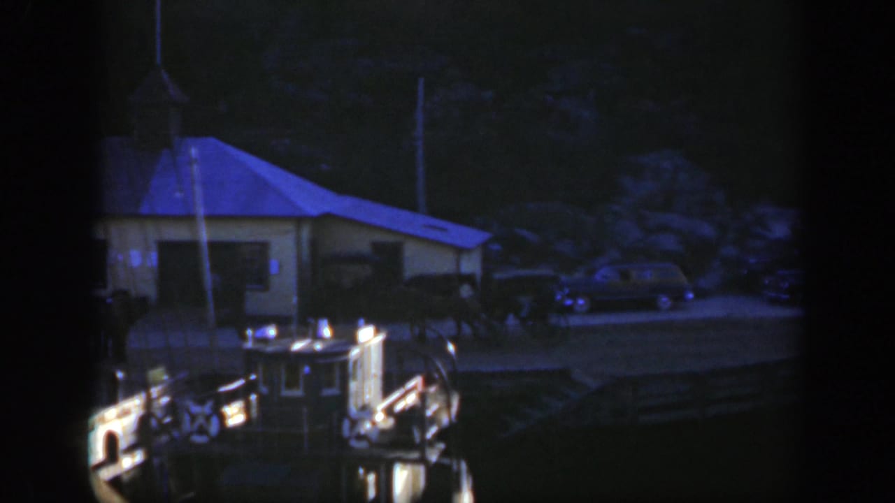 1950 Montreal waterfront view from a boat, with a small shoreline building, parked cars, dock railings, and deep blue low-light exposure.