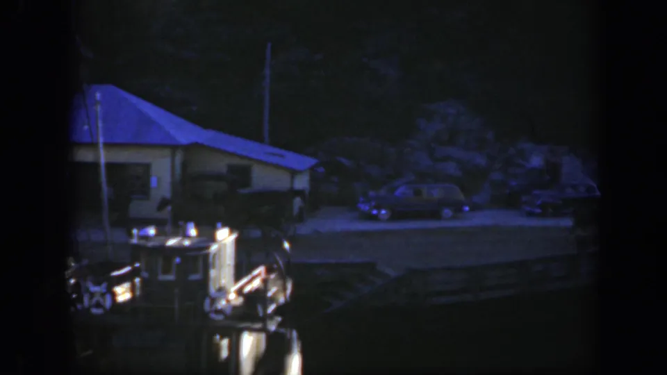 1950 Montreal home-movie frame looking from a boat toward a shoreline building, parked cars, and dark waterfront structures.