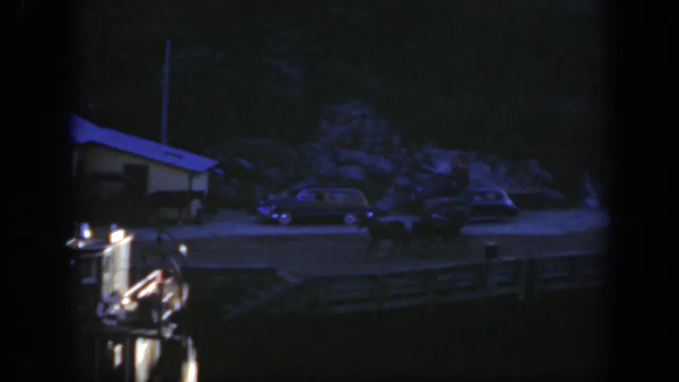 Dark 1950 Montreal dockside home-movie frame with vintage cars, a small waterside structure, and a possible horse-drawn cart or wagon.