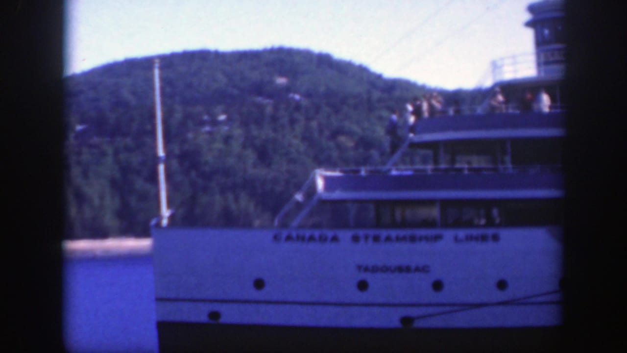 White Canada Steamship Lines vessel Tadoussac on the water near Montreal, with deck railings and a forested hill behind it.