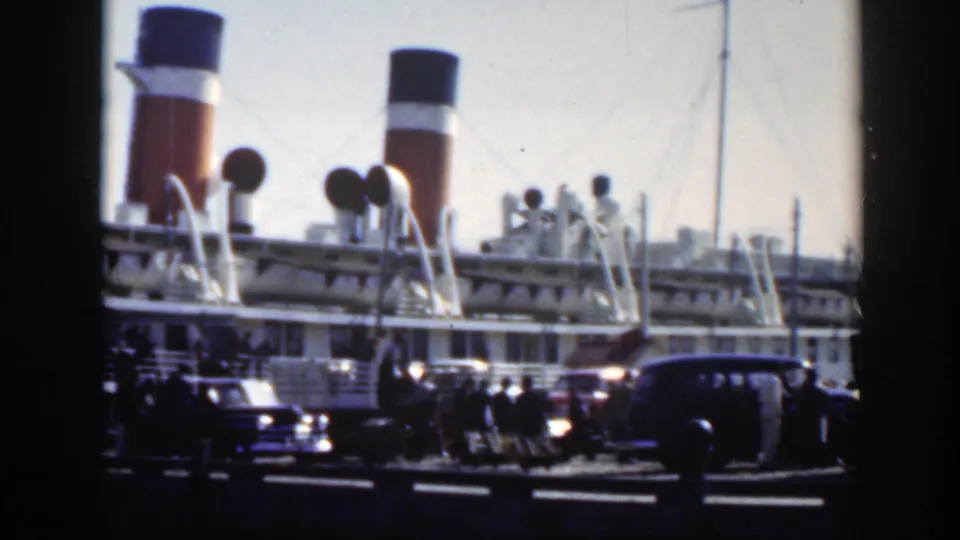 Dockside Montreal footage with the ship’s funnels and upper deck visible above parked mid-century cars and people at the pier.