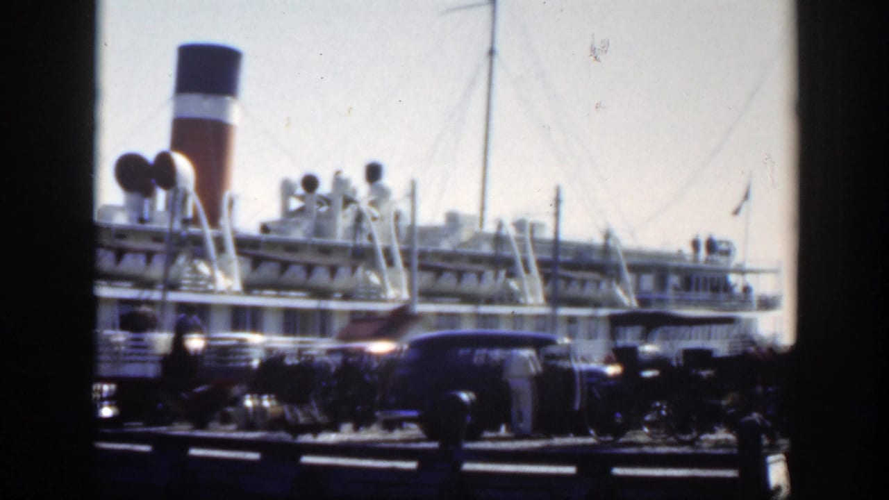 Dockside crowd and cars beside a large ship with a dark, white, and red funnel.