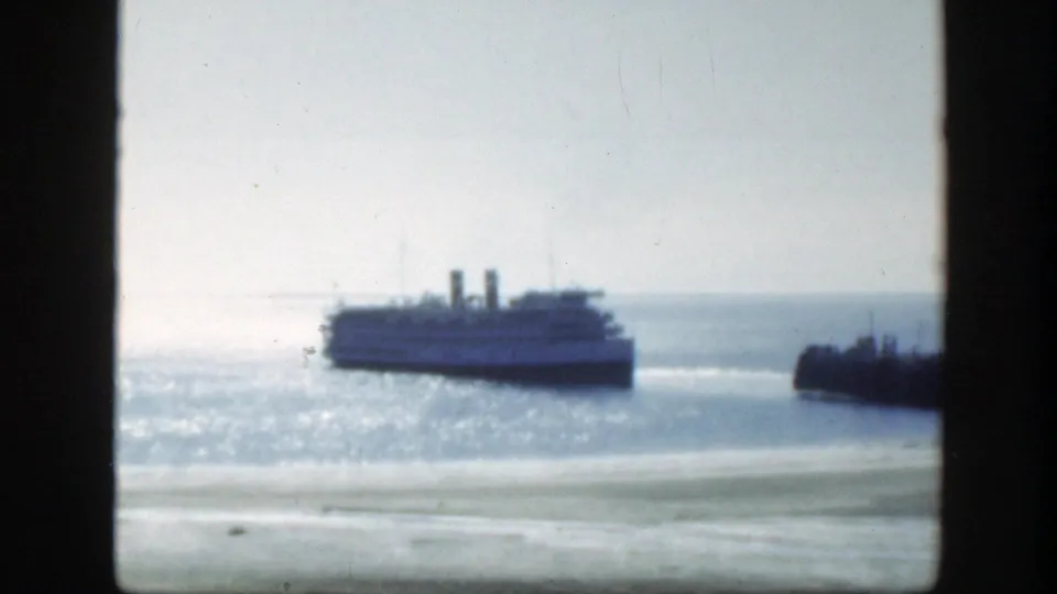 The liner is framed farther across the harbor, its two funnels visible against pale water and sky in the 8mm home-movie frame.
