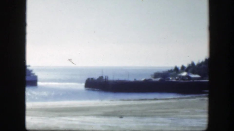 1950 8mm frame near Montreal showing bright water, a dark pier, shoreline trees, and part of a passenger vessel at frame edge.