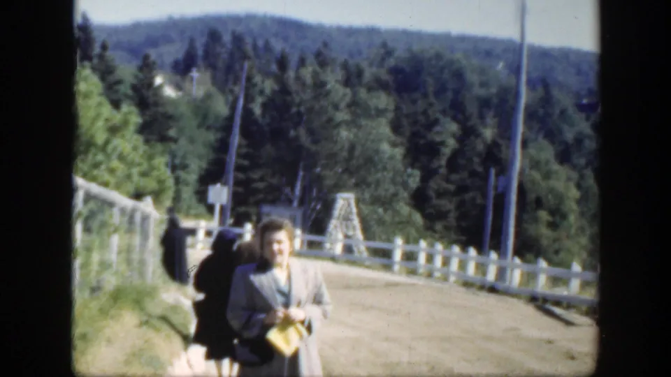 1950 8mm Montreal-area travel scene with an adult woman smiling on a rural road bordered by a white fence and evergreens.