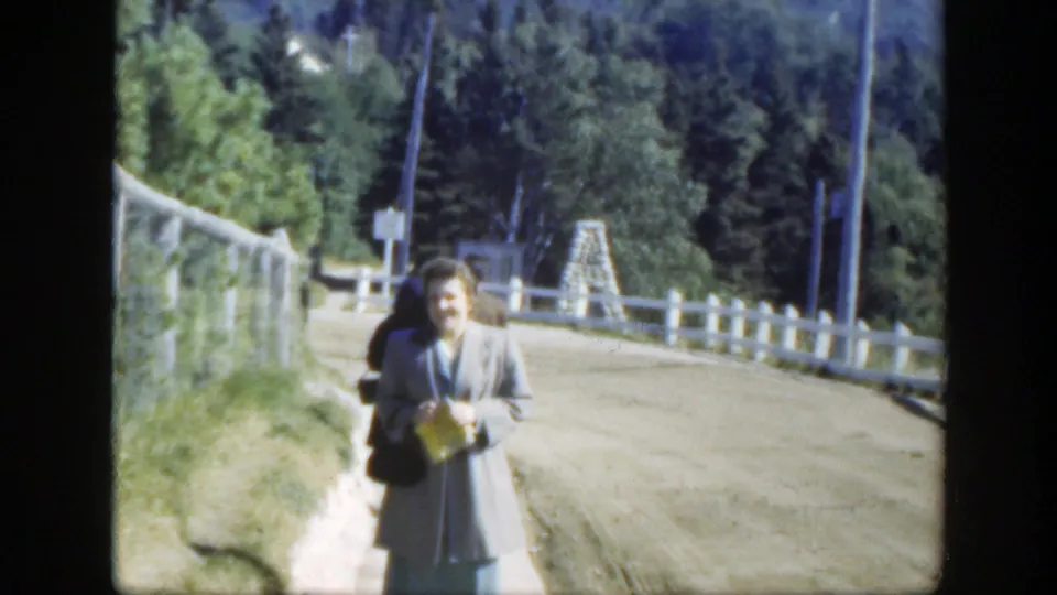 1950 8mm Montreal-area roadside view with a smiling adult woman, white fence, curving road, and wooded background.