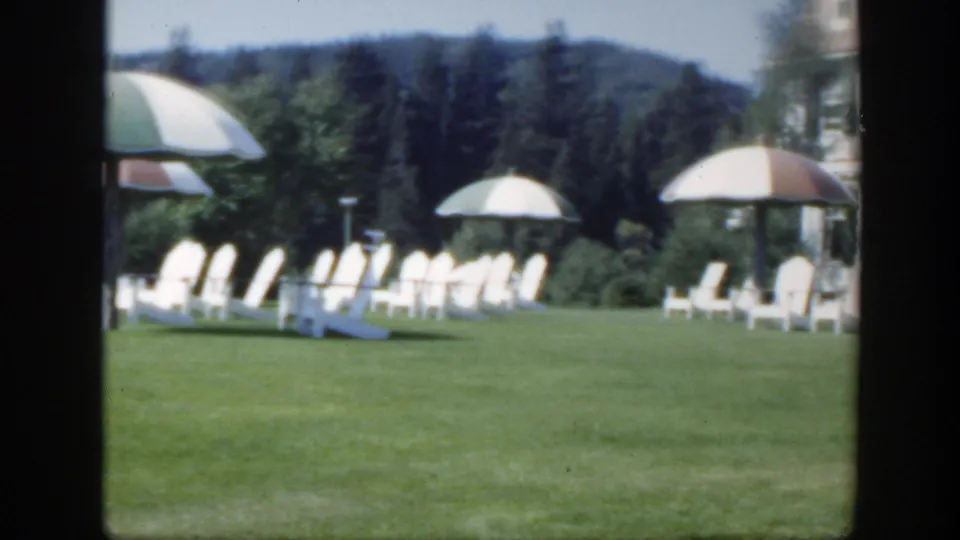 1950 Montreal home-movie view across a resort lawn toward white chairs, shade umbrellas, trees, and a wooded hill.