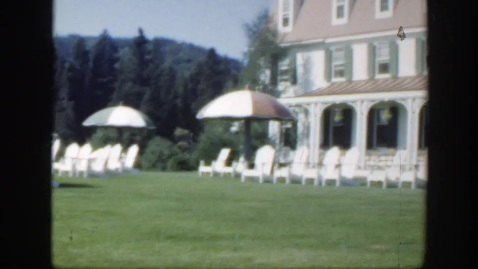 1950 Montreal resort exterior with a green-trimmed white house, lawn chairs, striped umbrellas, and wooded background.
