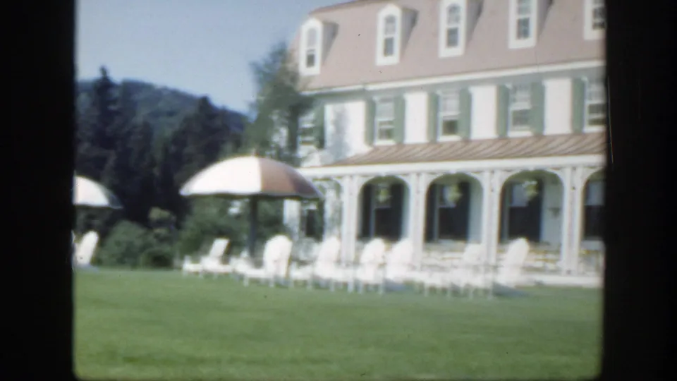 1950 Montreal home-movie frame showing a white hotel exterior with green shutters, veranda seating, lawn, and umbrellas.