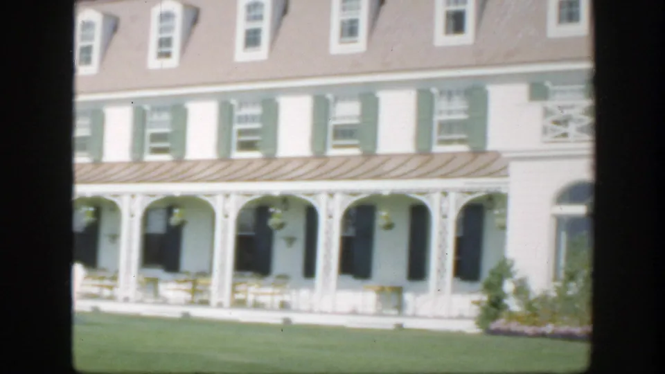 1950 Montreal home-movie frame showing a closer section of the hotel facade with veranda arches, shutters, lawn, and flowers.