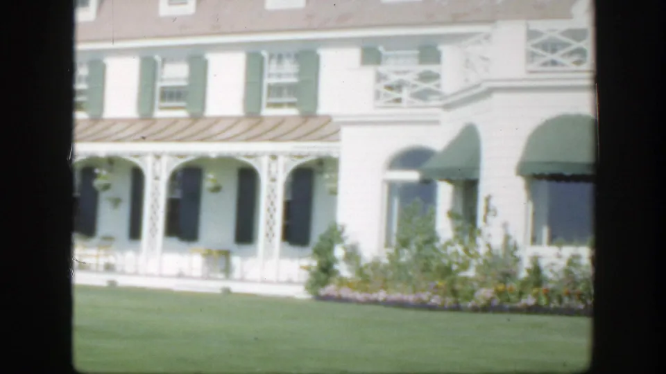 1950 Montreal home-movie view of the hotel entrance side, with green awnings, porch arches, shutters, lawn, and planted flowers.