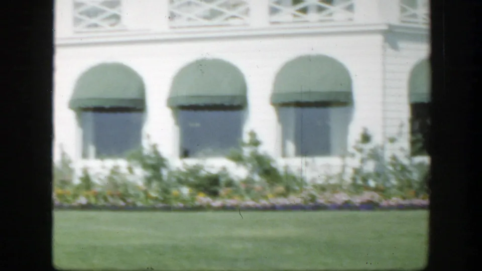 1950 Montreal home-movie frame with green awnings, white siding, flower beds, and a manicured lawn.