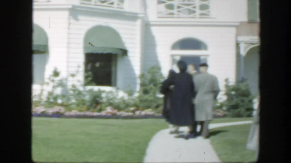 1950 Montreal 8mm frame of an elderly couple walking away on a path toward a white building with green awnings.