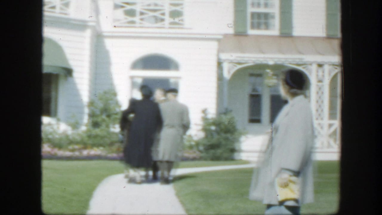 1950 Montreal home-movie frame showing adults in period coats near a white building with green shutters and a garden path.