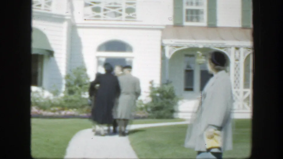 1950 Montreal home-movie frame showing adults in period coats near a white building with green shutters and a garden path.