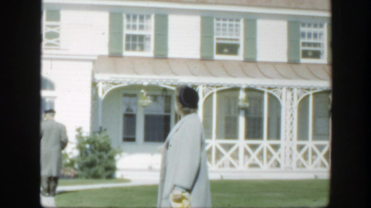 A woman in a light coat stands in the foreground while adults walk near a white porch.